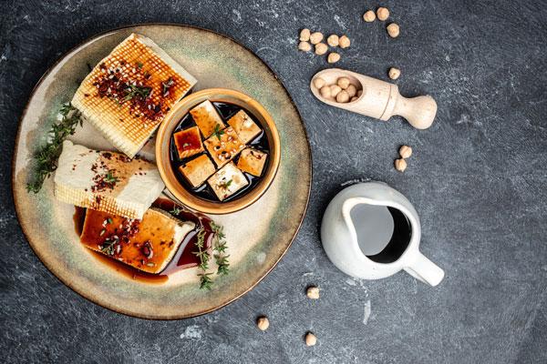 Image of marinated bean curd on a green ceramic plate sitting on a grey table with soy sauce and soybeans