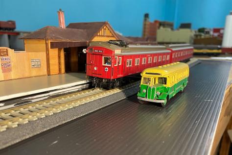 Toy red train on tracks parked at a station with yellow and green bus next to it