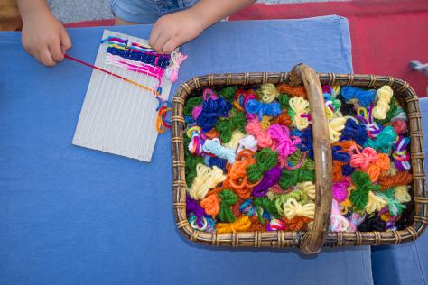 child weaving wool on a loom next to a basket full of wool