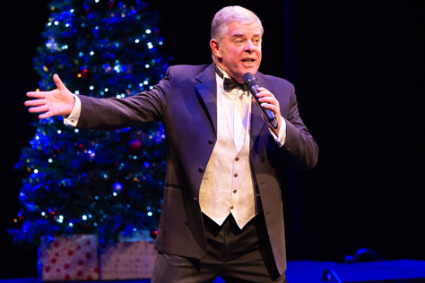 A man sings big in front of a lovely Christmas tree.