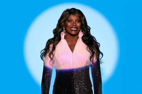 A woman, Marcia Hines, stands in a spotlight wearing a pink glitter top with long brown wavy hair flowing over her shoulders