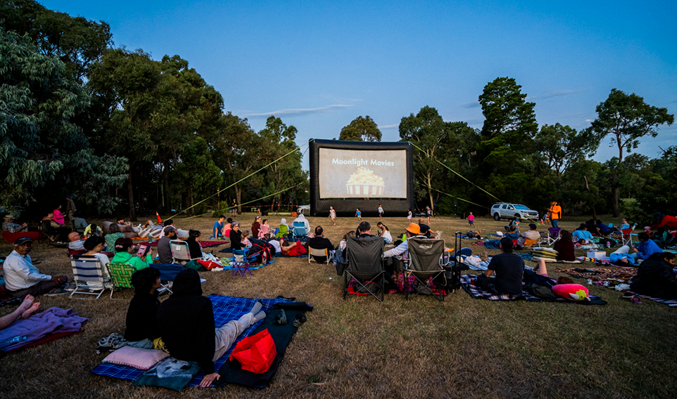 A wide shot of people seated on the grass waiting for an outdoor movie