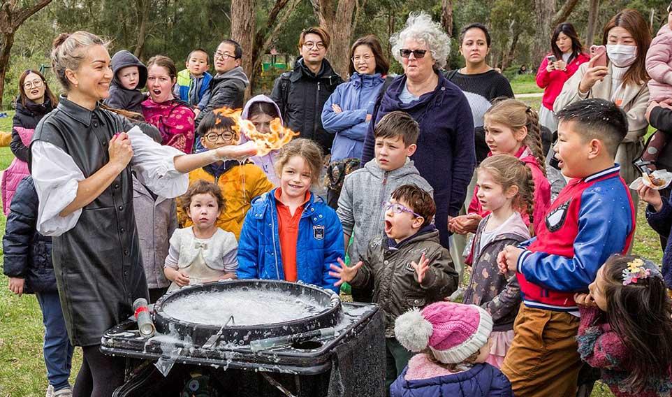 Group of children look on in amazement at woman doing a demonstration with flames in her hand